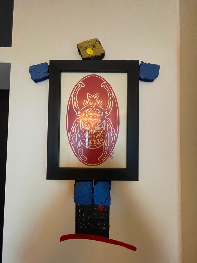 A colorful mixed-media art piece featuring a framed illustration of a beetle at the center, surrounded by abstract shapes and textures, including blue and yellow blocks at the top and a red curved line at the bottom, mounted on a light wall.